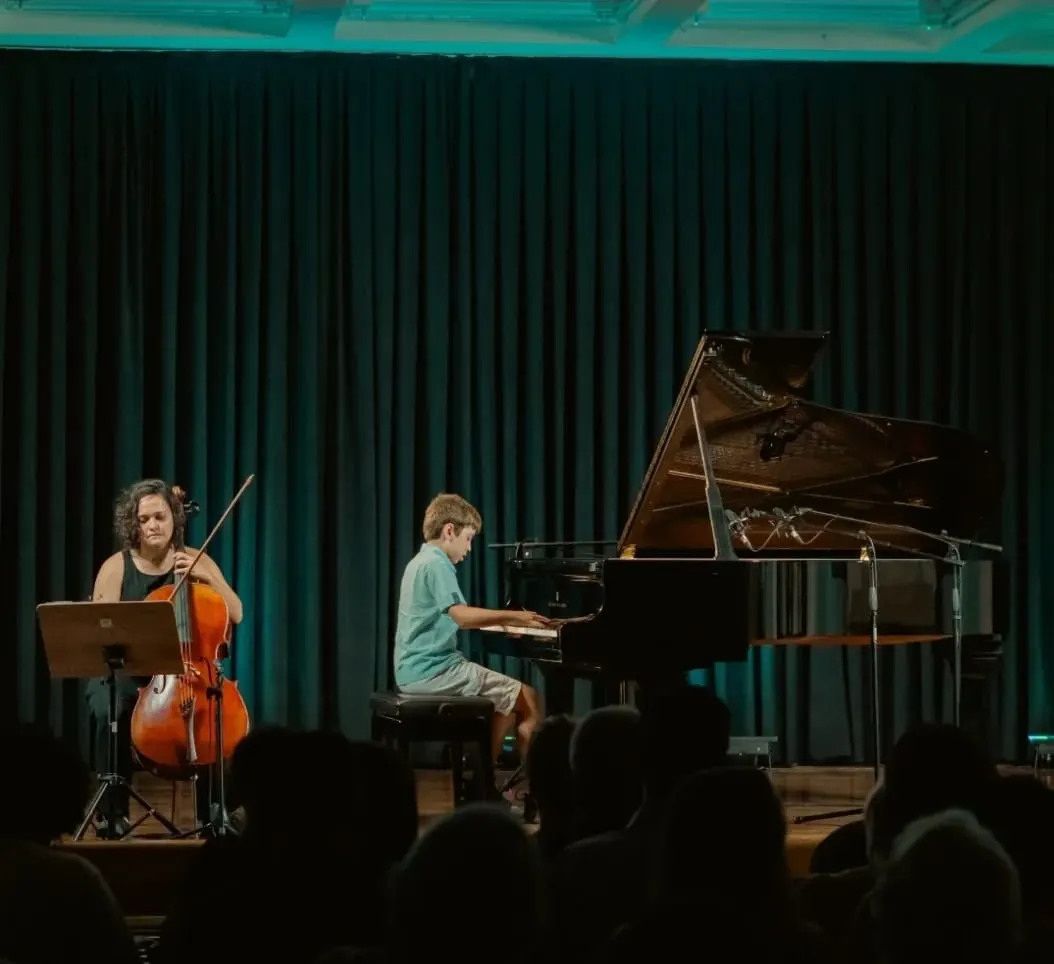 Aluno ao piano no palco com violoncelista